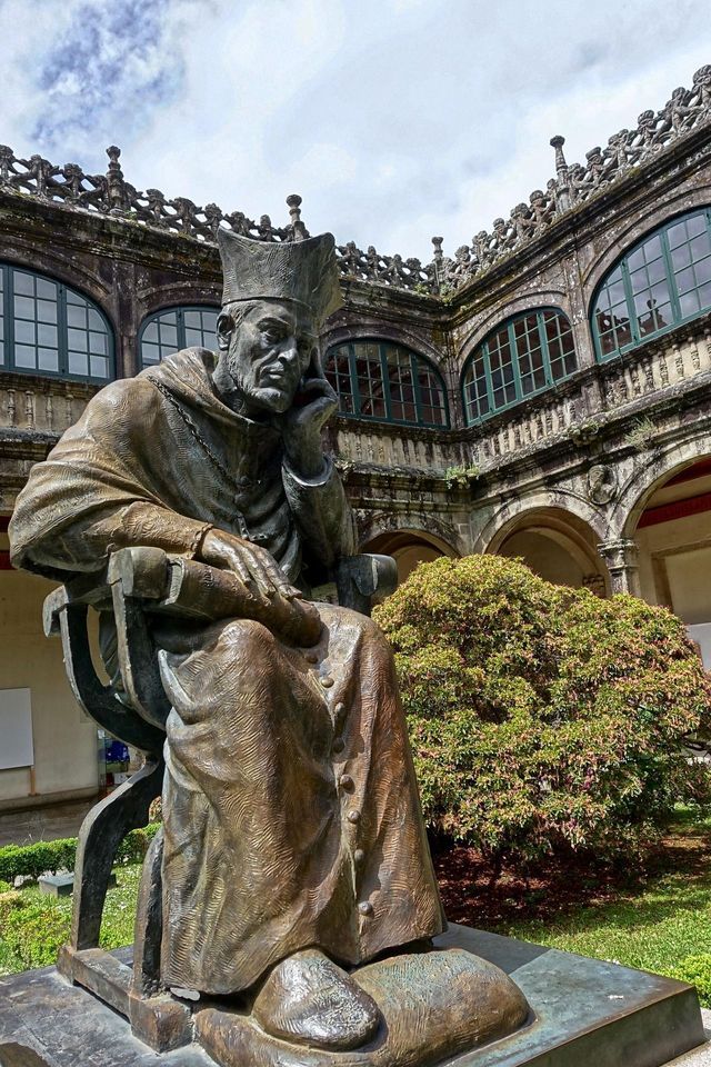 Una grande statua in bronzo di un chierico seduto in posa pensierosa, situata in un cortile storico in pietra con finestre ad arco.