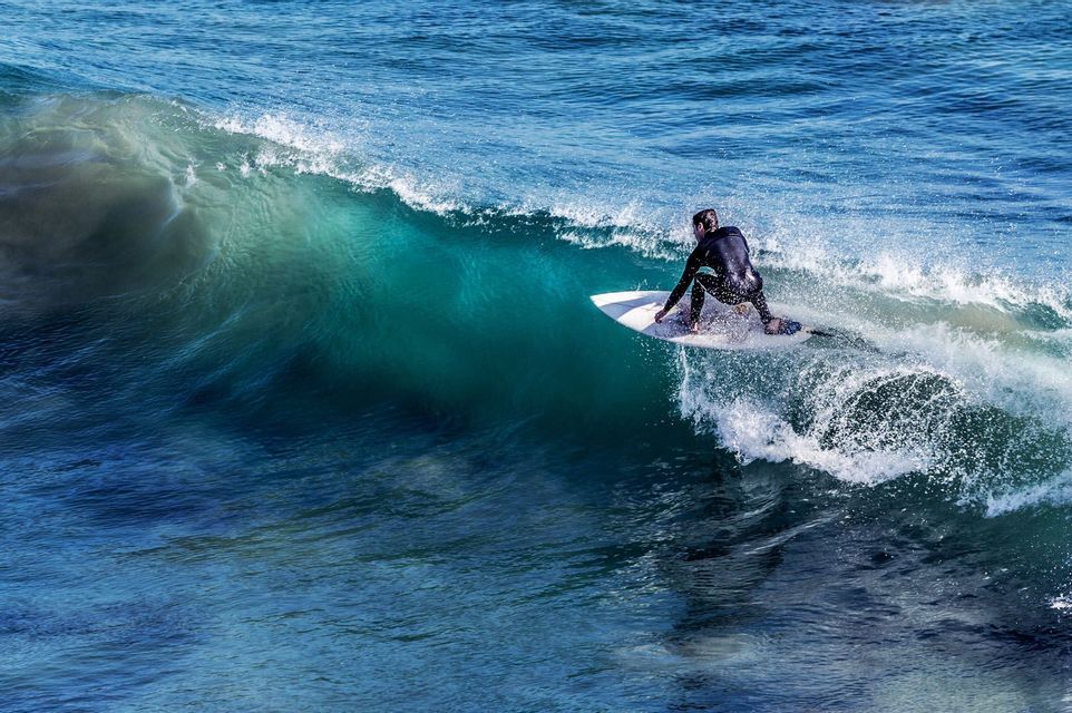Un surfista in muta nera cavalca la cresta di un'onda turchese che si increspa su una tavola da surf bianca, visto da una prospettiva aerea.