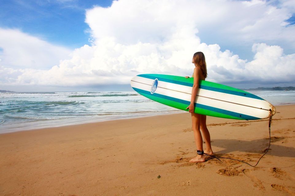 Una donna con la tavola da surf in piedi su una spiaggia di sabbia osserva le onde dell'oceano sotto un cielo parzialmente nuvoloso.