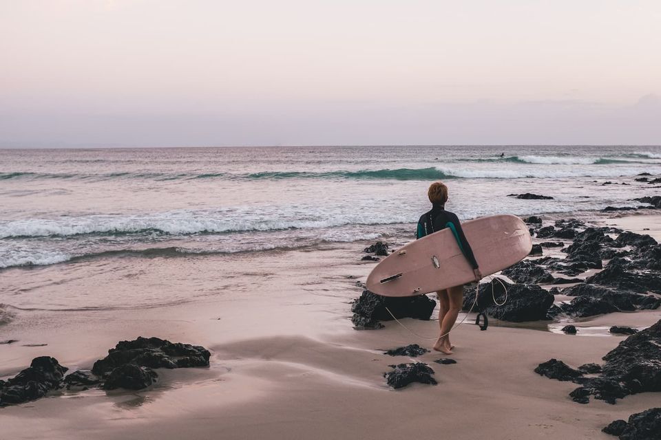 Un surfista con traje de neopreno lleva una tabla de surf rosa, caminando por una playa de arena y rocas hacia el océano al anochecer.