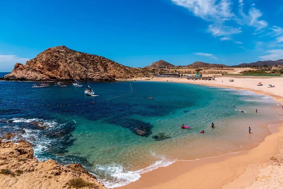 Una caletta soleggiata con una spiaggia sabbiosa, acqua cristallina color turchese, nuotatori e diverse barche ancorate nella baia.