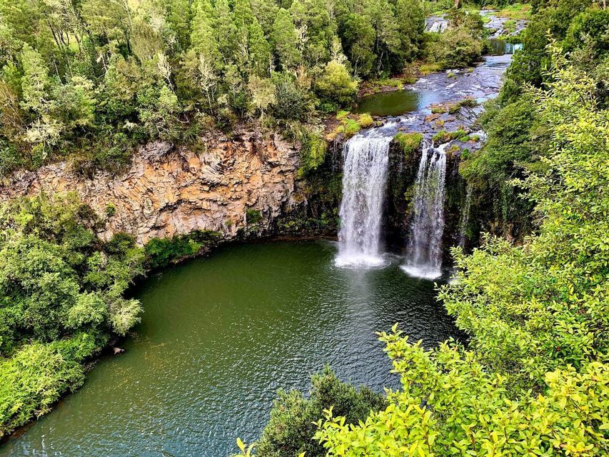 Vue aérienne d'une double cascade dévalant une falaise rocheuse dans une piscine verte, entourée d'une forêt dense.