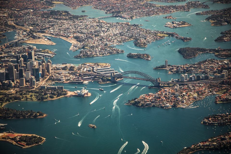 Une vue aérienne du port de Sydney, mettant en vedette l'Opéra et le Pont du Port, avec de nombreux bateaux naviguant sur les eaux bleues.