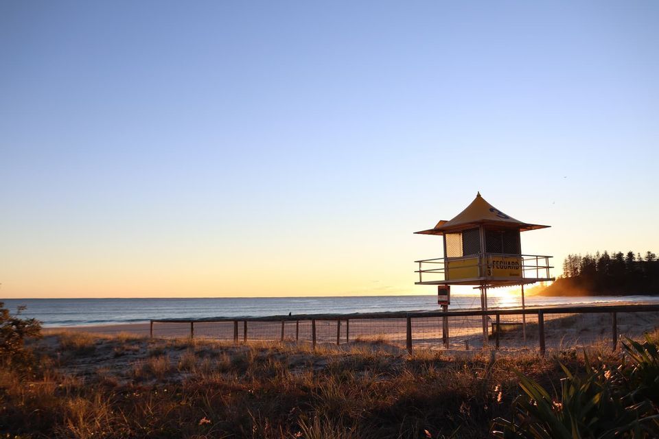 Una torre de salvavidas amarilla se alza en una playa de arena al amanecer, con vistas al océano en calma bajo un cielo despejado.