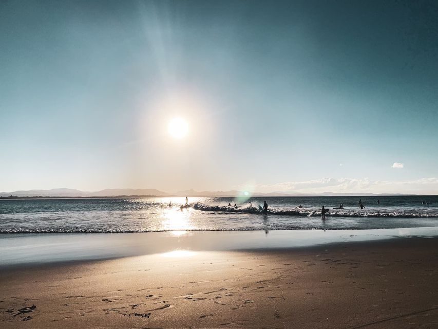 Surfistas en el océano cabalgando olas al atardecer, vistos desde una playa de arena con el sol reflejado en el agua.