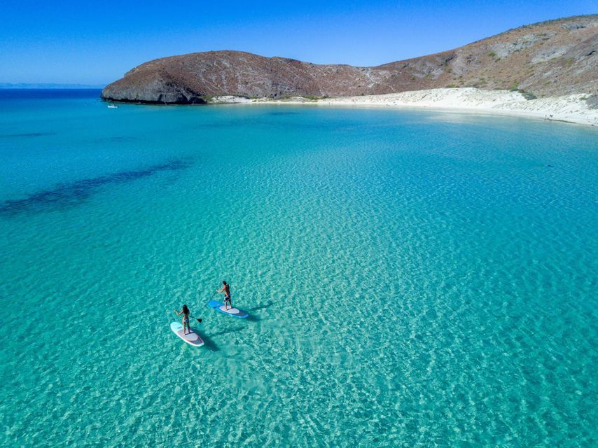 Veduta aerea di due persone su paddleboard in una baia con acque cristalline turchesi e una spiaggia di sabbia bianca.