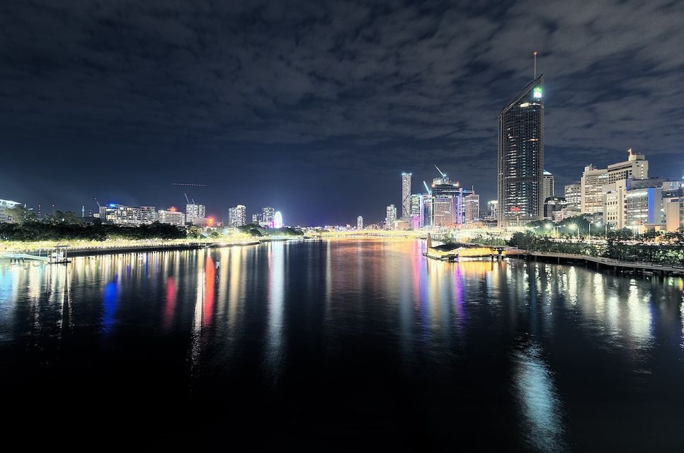 Un large fleuve reflète les lumières colorées d'une ligne d'horizon urbaine moderne et de hauts bâtiments sous un ciel de nuit nuageux.