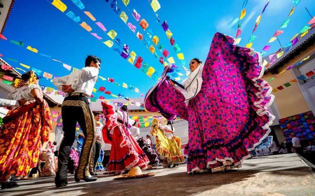 Ballerini in vivaci costumi tradizionali si esibiscono su un palco all'aperto sotto bandiere colorate di festival contro un cielo blu.
