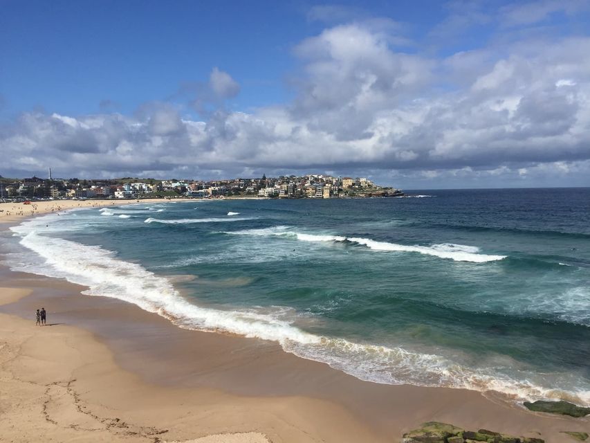 Un pueblo costero domina una amplia playa de arena mientras las olas turquesas rompen en la orilla bajo un cielo parcialmente nublado.