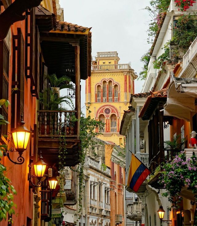 Una strada stretta è fiancheggiata da colorati edifici coloniali con balconi in legno, lampioni accesi e una bandiera colombiana.