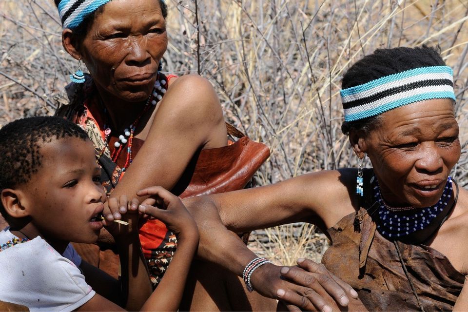 Zwei indigene Frauen und ein kleines Kind sitzen draußen zusammen und tragen traditionelle Kleidung und Perlenschmuck.