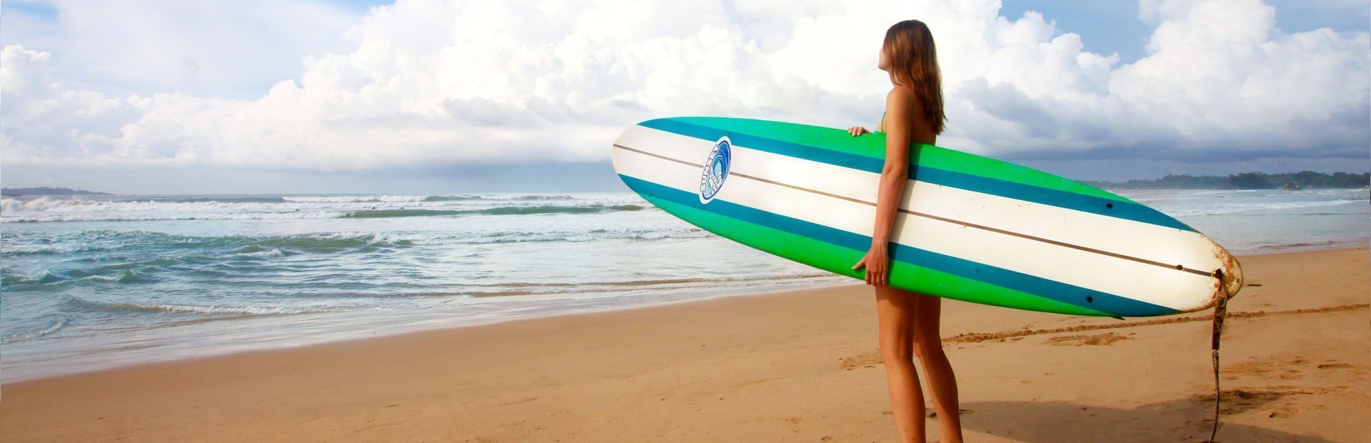 Una donna con una tavola da surf verde e bianca si trova su una spiaggia sabbiosa, guardando l'oceano sotto un cielo nuvoloso.