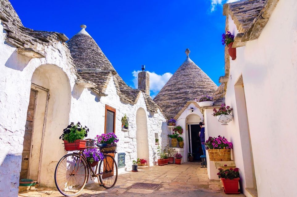 A vintage bicycle with baskets of purple flowers is parked on a stone alley between whitewashed houses with conical roofs under a blue sky.