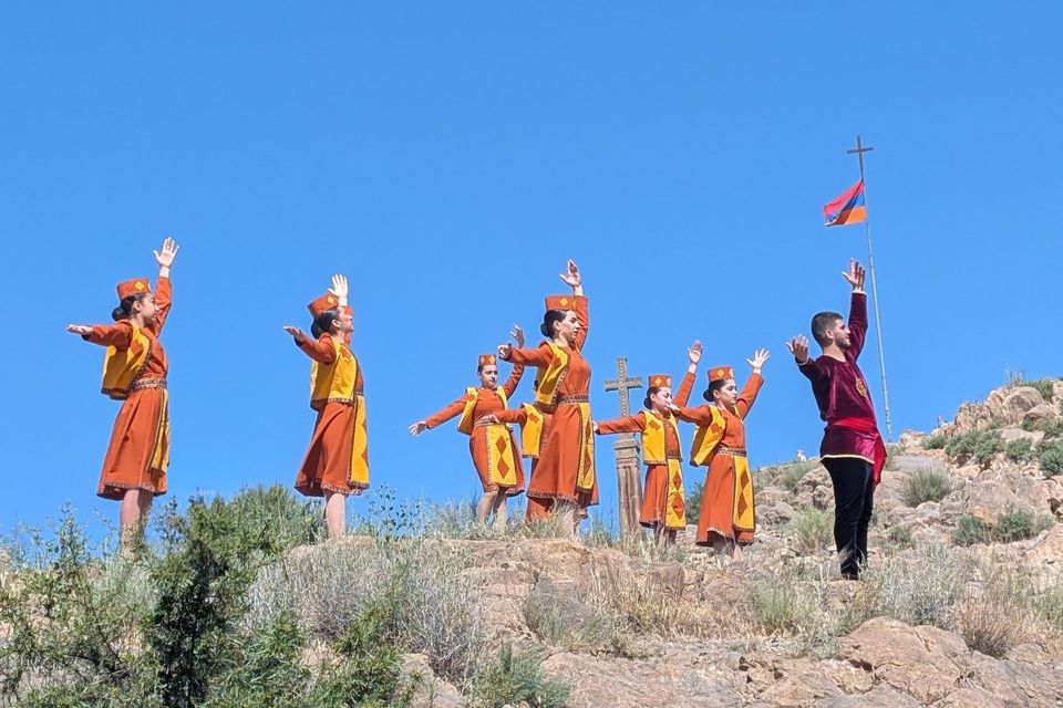 Eine Gruppe von Personen in traditionellen orange-gelben Kostümen vollführt einen synchronisierten Tanz auf einem felsigen Berghang unter strahlend blauem Himmel, neben einer armenischen Flagge.
