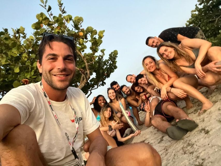 Ein Mann macht ein Selfie mit einer großen, lächelnden WeRoad-Gruppe an einem Sandstrand unter klarem Himmel.