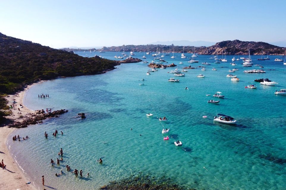 Una vista aerea di una baia affollata con acqua turchese cristallina, dove le persone nuotano vicino alla riva e numerose barche sono ancorate.