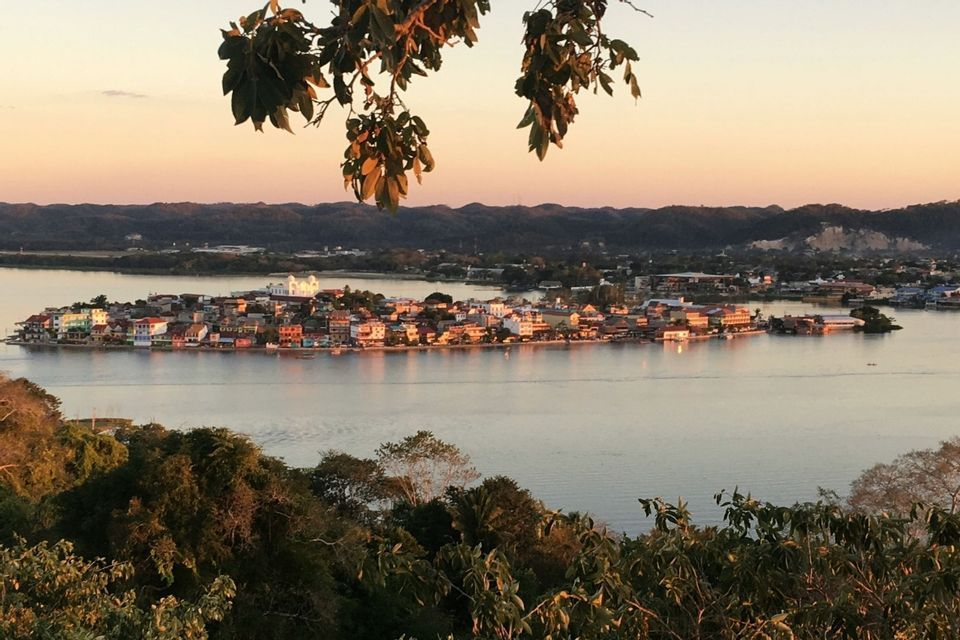 Eine Vogelperspektive einer farbenfrohen Inselstadt an einem ruhigen See bei Sonnenuntergang, umrahmt von Baumblättern im Vordergrund.