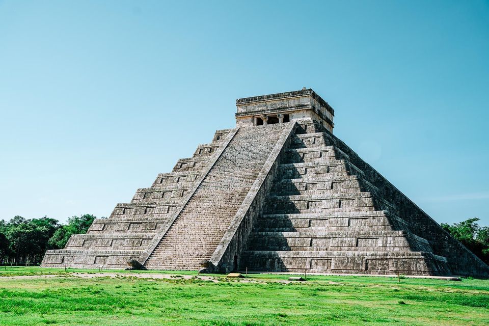 Eine große steinerne Stufenpyramide mit einer zentralen Treppe steht auf einem grünen Rasen unter einem klaren blauen Himmel.