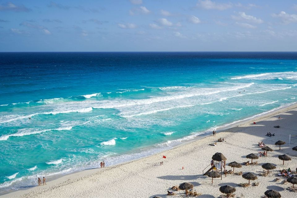 Luftaufnahme eines weißen Sandstrandes mit türkisfarbenen Wellen, gesprenkelt mit Strohschirmen und sonnenbadenden Menschen.