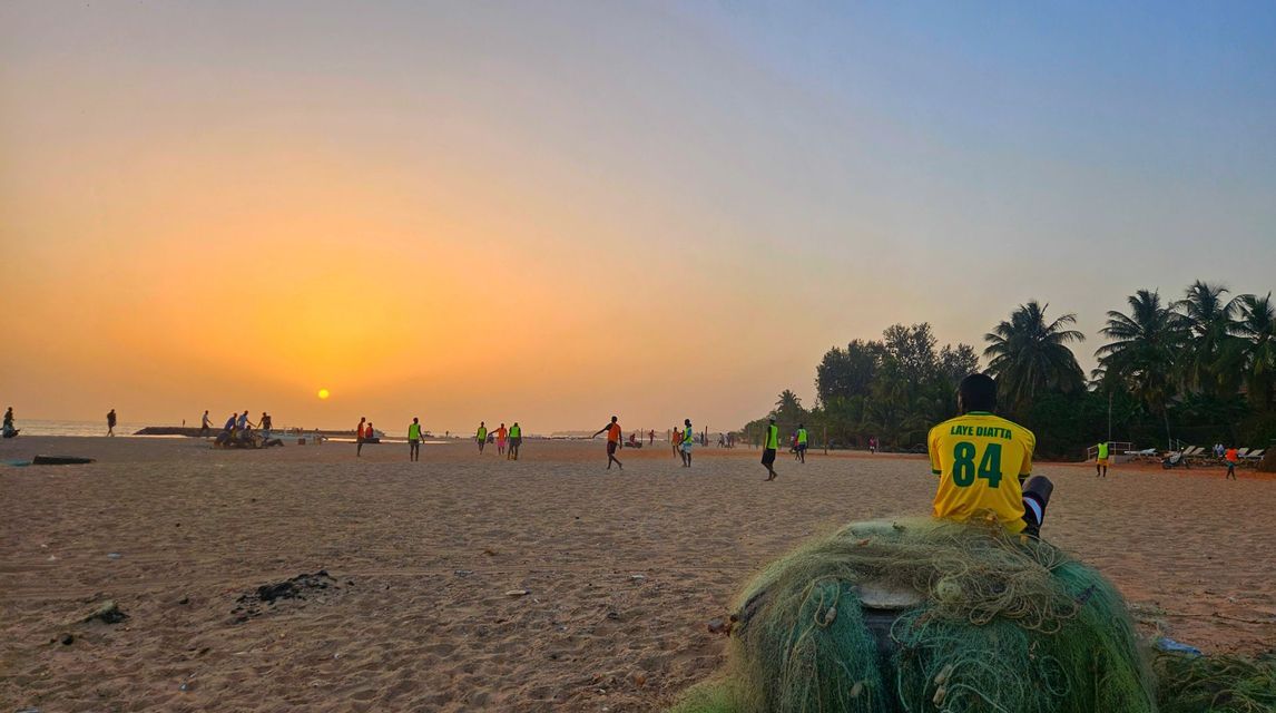 Eine Person in einem gelben Trikot sitzt bei Sonnenuntergang auf Fischernetzen und beobachtet Menschen beim Fußballspielen am Sandstrand.