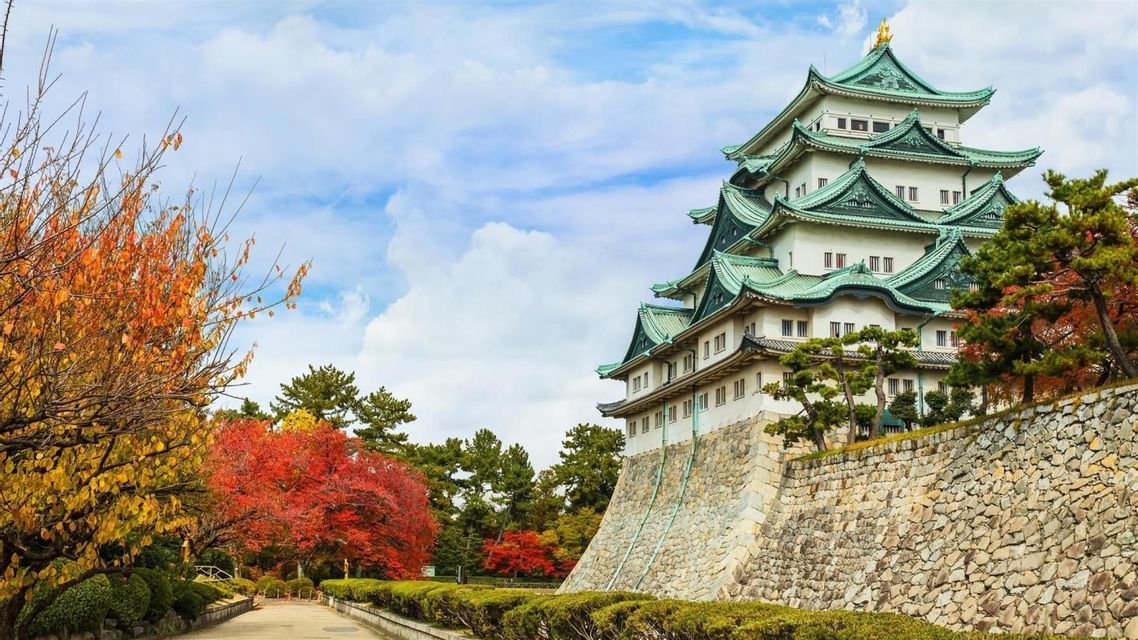 Un tradizionale castello giapponese con tetti verdi si erge su un muro di pietra, circondato da alberi con fogliame autunnale colorato sotto un cielo nuvoloso.