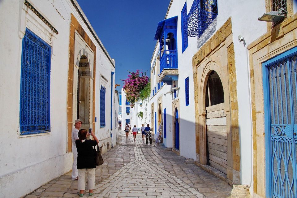 Persone camminano lungo una stretta strada acciottolata, fiancheggiata da edifici bianchi con porte e finestre blu brillante, sotto un cielo sereno.