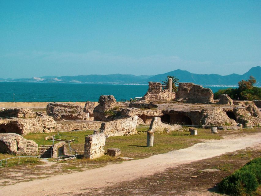 Antiche rovine in pietra su un appezzamento erboso in riva al mare, con montagne lontane visibili oltre l'acqua sotto un cielo sereno.