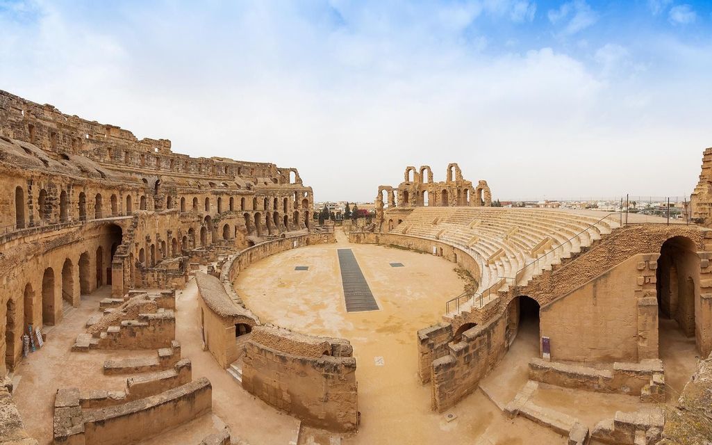 Una vista panorámica de las ruinas de un gran anfiteatro antiguo con asientos de piedra escalonados y una arena central arenosa.
