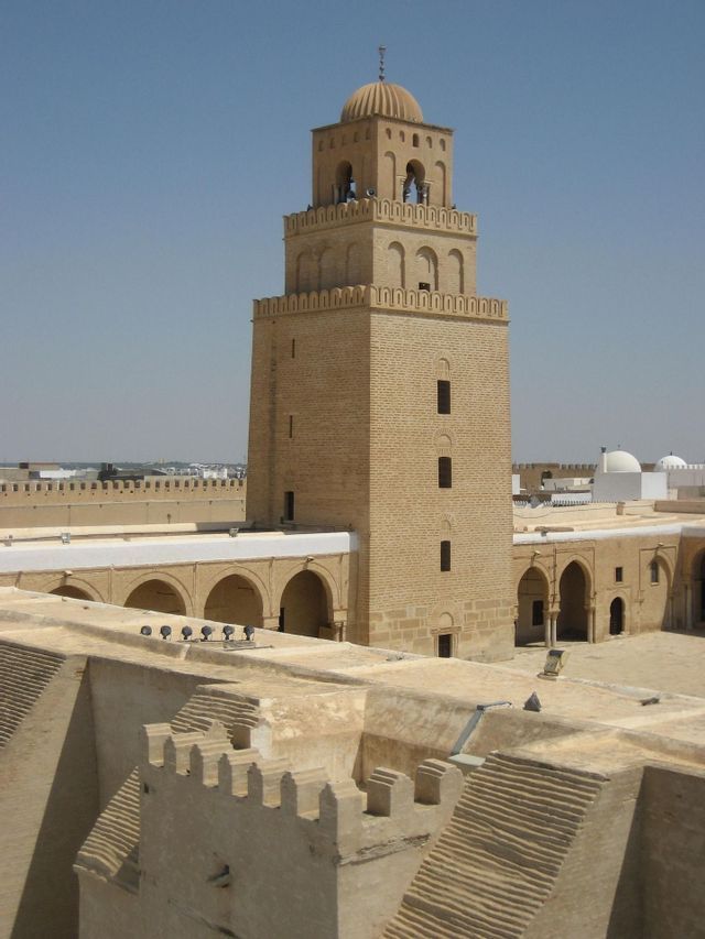 Un minarete cuadrado de piedra color arena con una cúpula se alza sobre los tejados de un antiguo complejo bajo un cielo azul claro.