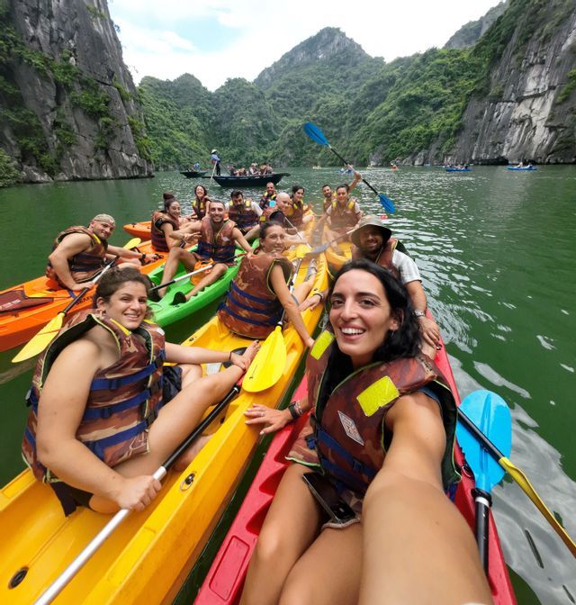 Una mujer se toma un selfie sonriente durante un viaje en grupo de WeRoad, en kayaks coloridos sobre aguas verdes y rodeada de acantilados escarpados y exuberantes.