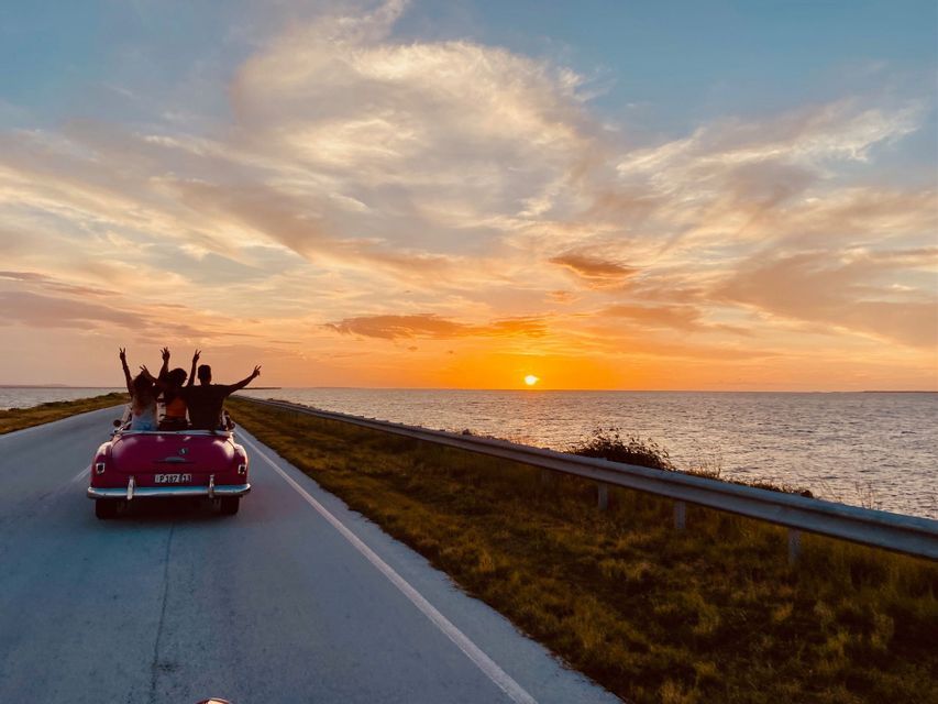 Un viaggio di gruppo WeRoad visto da dietro, alla guida di una decappottabile vintage rosa su una strada costiera durante un tramonto dorato.