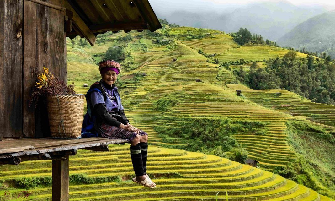 An elderly woman in traditional clothing sits smiling on a wooden porch overlooking vast, terraced rice paddies on a hillside.