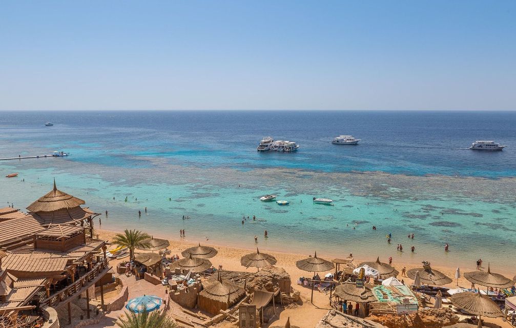 Una vista elevada de una playa de arena con sombrillas de paja, personas nadando en aguas turquesas y varios yates amarrados en el mar.