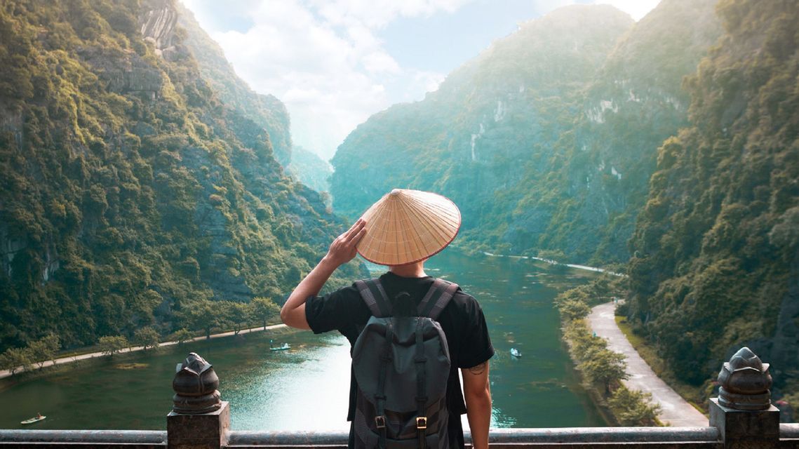 A person with a backpack and a conical hat stands on a viewpoint overlooking a river valley surrounded by lush green mountains.