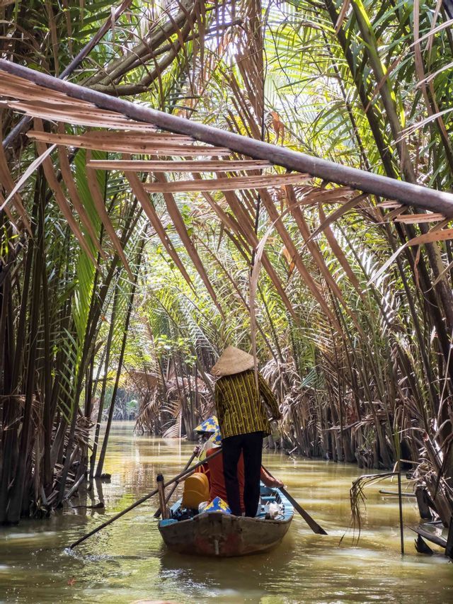 A WeRoad group trip rows a boat down a narrow river, navigating through a dense tropical palm forest.