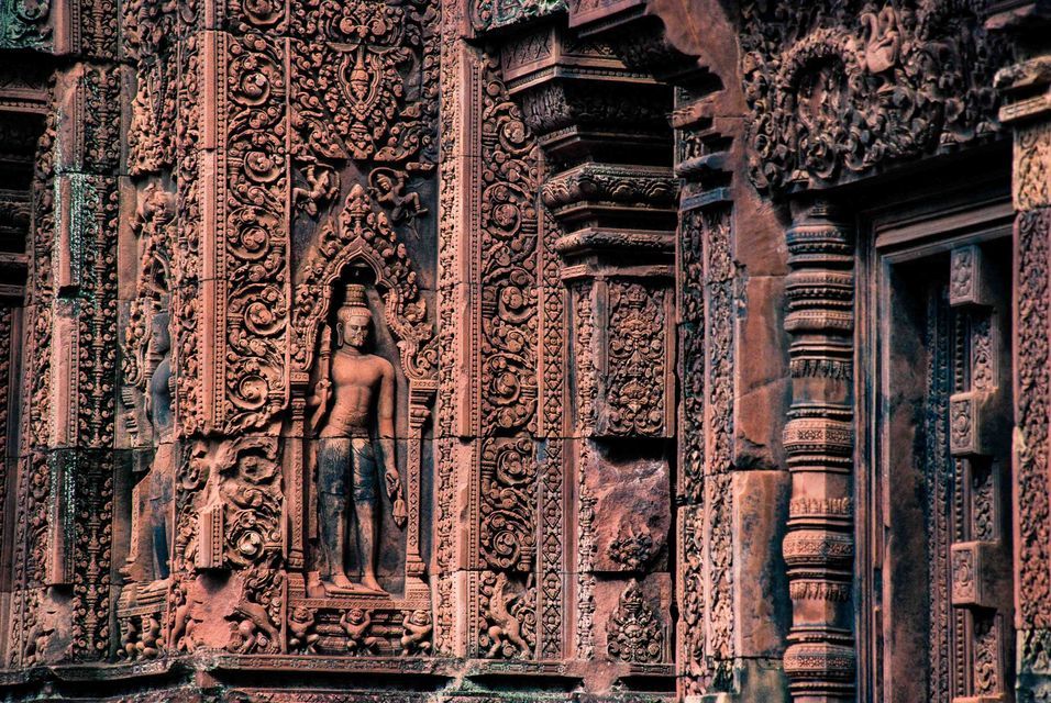 Close-up of intricate carvings on a reddish stone temple wall, featuring a standing deity in a decorative niche.