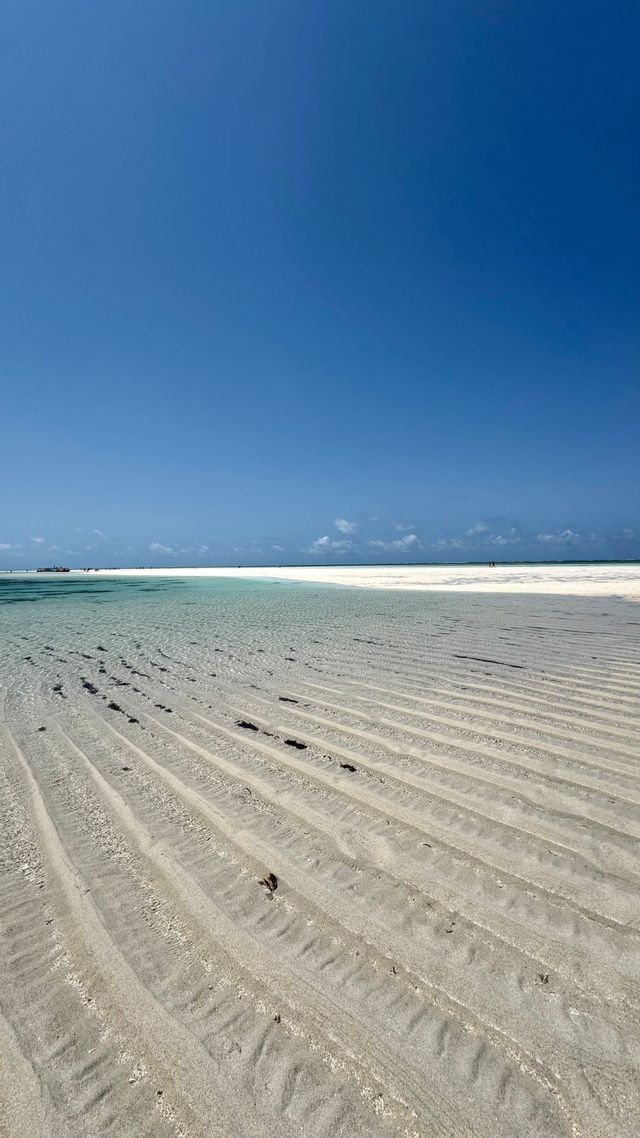 Patrones de arena ondulada en agua clara y poco profunda en una playa de arena blanca bajo un cielo azul vasto y sin nubes.