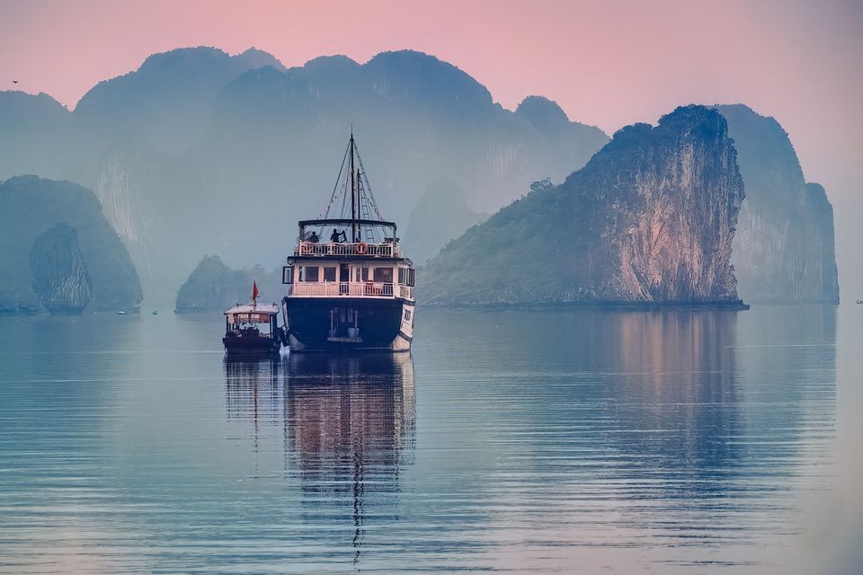 Un voyage en groupe WeRoad sur un bateau de tourisme flottant sur une eau calme au milieu de karsts calcaires brumeux au crépuscule rose.