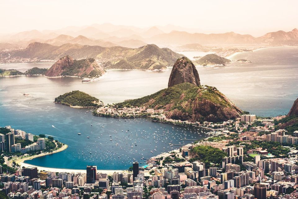 An aerial view of a sprawling coastal city with a large mountain overlooking a bay filled with boats, with hazy hills in the distance.