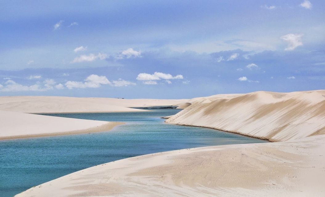 A vibrant blue lagoon winds through expansive white sand dunes under a sky dotted with fluffy white clouds.