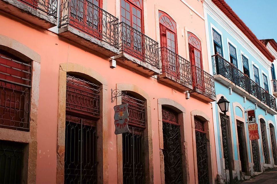 Un edificio coloniale rosa con finestre rosse si trova accanto a uno blu, entrambi con balconi in ferro battuto.