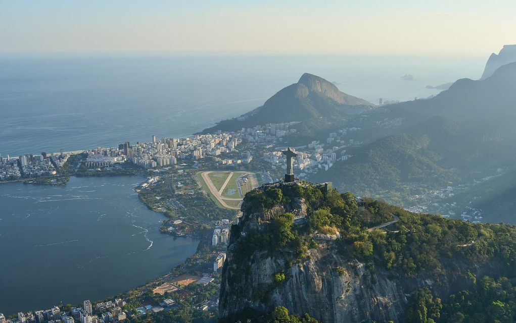 An aerial view of the Christ the Redeemer statue on a mountain, overlooking a coastal city with a large lagoon and the ocean.