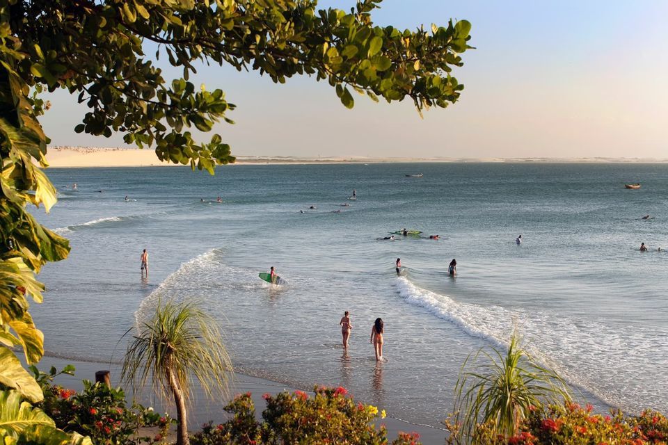 A high-angle view of people surfing and swimming in the ocean at a beach, with sand dunes visible in the distance.