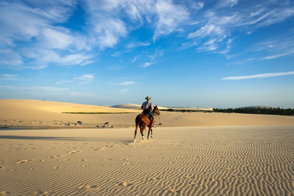 A person wearing a hat rides a brown horse across vast, rippled sand dunes under a bright blue sky with wispy clouds.