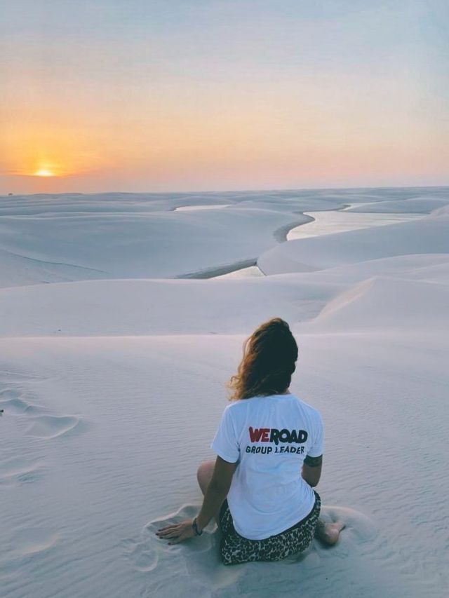 Un capogruppo WeRoad siede su una duna di sabbia bianca, guardando il tramonto su un paesaggio di lagune serpeggianti.