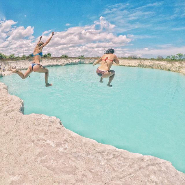 Two women in bikinis jumping from a rocky ledge into a pool of turquoise water under a blue, cloudy sky.