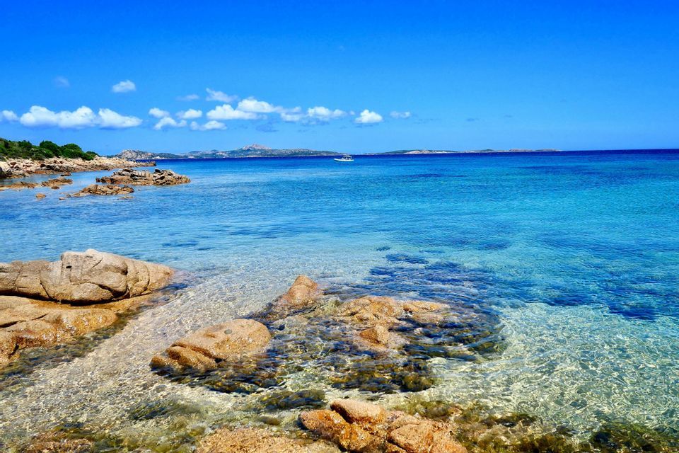 Una costa rocciosa incontra acque calme, limpide e turchesi sotto un cielo azzurro brillante con una singola barca in lontananza.