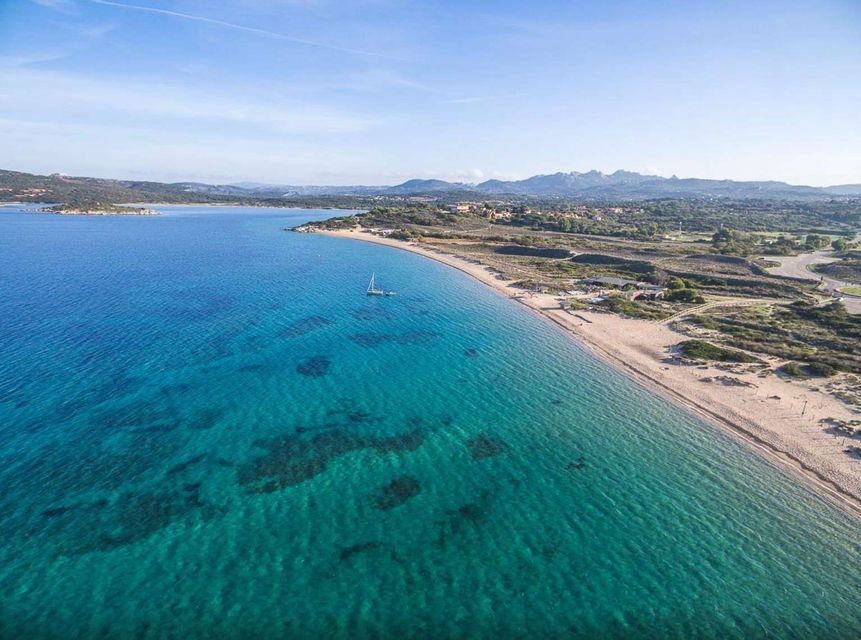 Vista aerea di una barca a vela solitaria su acqua cristallina turchese lungo una spiaggia sabbiosa, con colline e montagne verdi in lontananza.