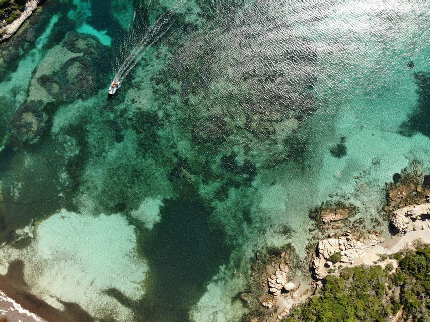 Veduta aerea di una piccola barca che naviga su acque turchesi limpide, lasciando una scia, vicino a una costa rocciosa.