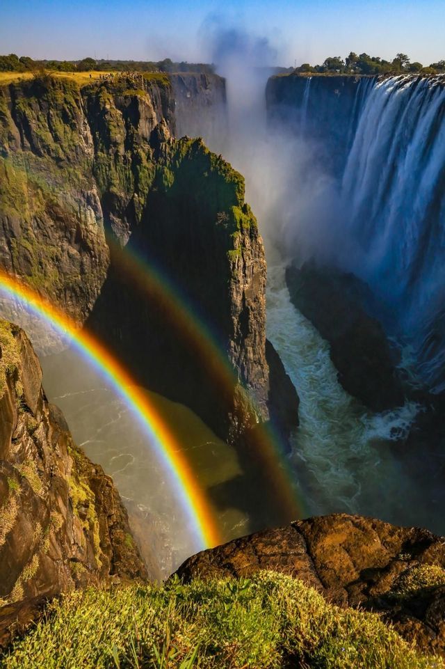 Un doppio arcobaleno si inarca su una gola profonda e rocciosa con una grande cascata sullo sfondo sotto un cielo sereno.